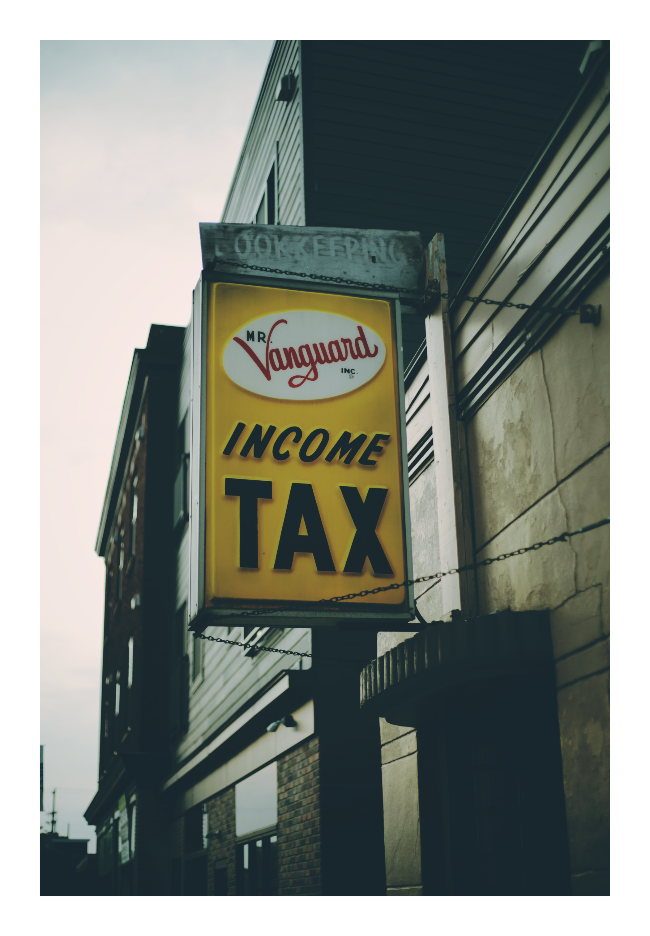 A vertically oriented photograph of a bright yellow “INCOME TAX” sign with red and black lettering hanging from the side of a building by metal chains. The sign is mounted beneath a weathered wooden panel and set against the exterior of older buildings at dusk.